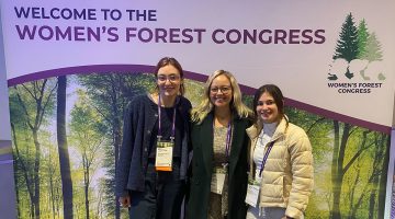 Three female students wearing conference badges stand in front of a sign that says “Welcome to the Women’s Forest Congress.”