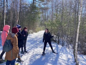 Several people in snowshoes are gathered around graduate student Colby, who has one arm extended to point out a tree. A forested trail covered in snow stretches out into the background, dappled with the shadows of trees.